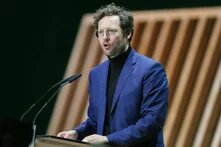 Ein Mann mit Brille und lockigem Haar spricht an einem Rednerpult mit zwei Mikrofonen, im Hintergrund schräge Holzlamellen.