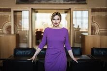 Person mit blonden Haaren und violettem Kleid steht vor einem Tisch, stützt sich mit beiden Händen ab, schaut in die Kamera; im Hintergrund geöffnete Tür und Besprechungsraum.