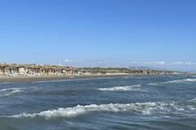 Waves hit a wide sandy beach with many loungers under thatched roofs, trees and mountains in the background.