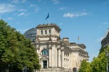Reichstagsgebäude in Berlin mit Glaskuppel, deutscher und EU-Flagge, teils von Bäumen verdeckt.