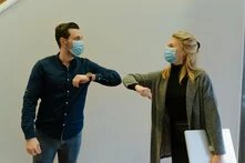 Zwei Personen mit Maske begrüßen sich mit Ellbogenkontakt in Innenraum, eine hält einen Laptop