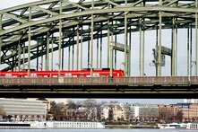 Rote Regionalbahn fährt über die Hohenzollernbrücke in Köln, darunter der Rhein mit zwei weißen Schiffen.