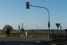 Rote Ampel an Landstraße mit Blick auf Felder, Strommasten und Verkehrsschilder bei klarem Himmel.