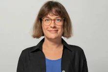 Person mit kinnlangem braunem Haar, Brille und dezentem Lächeln blickt frontal in die Kamera. Hintergrund ist einfarbig hell.