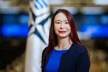 Frau mit langen Haaren steht vor unscharfer Flagge mit Sternmotiv, schaut in die Kamera, trägt dunkles Oberteil.