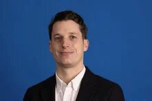 Person mit kurzem dunklem Haar und hellem Hemd vor blauem Hintergrund, schaut in die Kamera.