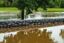 Ein Sandsackwall trennt braunes Hochwasser von überfluteter Wiese mit Baum im Hintergrund.
