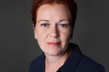 Das Bild zeigt das Porträt einer Frau mit kurzen rötlichen Haaren und hellem Teint in einem dunklen Oberteil vor einem neutralen, grauen Hintergrund.