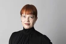 Person mit rotem Ponyblick und dunklem Oberteil vor grauem Hintergrund, schaut in die Kamera.