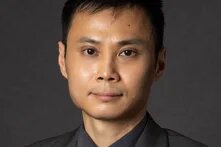 Person mit kurzem, dunklem Haar blickt frontal in die Kamera, trägt Anzug, Hemd und helle Krawatte vor dunklem Hintergrund.