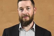 Eine Person mit Bart steht vor einer einfarbigen Wand und blickt in die Kamera.