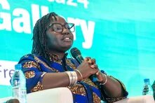 Fatou Warkha Sambe, wearing glasses and a patterned blue blouse, sits on a stage, holding a microphone and speaking in front of a turquoise screen.