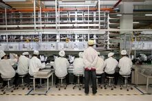 Neun Personen sitzen an Werkbänken, eine steht dahinter; alle tragen helle Kittel und Mützen in einer Fabrikumgebung.