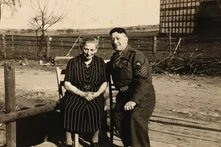 Zwei Personen sitzen draußen nebeneinander, eine ältere Person in Kleid, eine in Uniform; im Hintergrund Zaun und Felder.