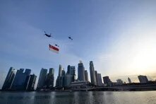 Drei Hubschrauber fliegen über Skyline, einer trägt die Flagge von Singapur, unten Wasser und Hochhäuser bei Tageslicht.