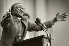 Der südafrikanische Geistliche Desmond Tutu steht am Rednerpult mit Mikrofonen, trägt Klerikalkragen und Kreuzkette, Arme weit geöffnet, lächelt ins Publikum.