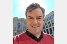 Person macht ein Selfie im Freien, lächelt in die Kamera, im Hintergrund Gebäude mit Fenstern und Figuren.