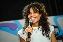 Person in weißem T-Shirt spricht in ein Mikrofon. Sie trägt dunkle, lange Locken, ein Nasenpiercing und pinken Lippenstift
