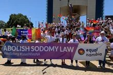 Many people stand in front of a monument on a staircase with rainbow flags and a banner from the organization Equal Namibia that reads “ABOLISH SODOMY LAW.”