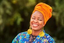 Lächelnde Person mit orangefarbenem Kopftuch und großen runden Ohrringen, vor unscharfem grünem Hintergrund.