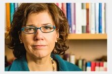 Porträt einer Frau mit Brille, frontal, ernster Blick. Im Hintergrund unscharfe Bücherregale.