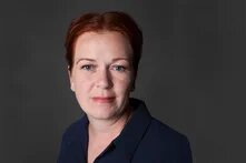 Porträt einer Frau mit kurzen Haaren, frontal, neutraler Blick. Sie trägt ein Oberteil, Hintergrund dunkel und einfarbig.