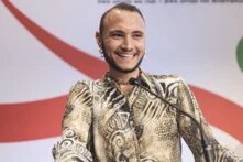 Eine lächelnde Person mit kurzen dunklen Haaren und Bart steht an einem Rednerpult mit Mikrofon, über dem eine Regenbogenflagge hängt.