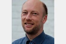 Porträt eines lächelnden Mannes mit Glatze und Bart, frontal aufgenommen. Er trägt Hemd und Krawatte, Hintergrund unscharf.