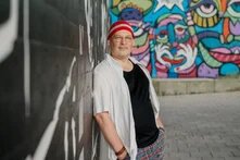 Mensch mit grauen Bartstoppeln, roter Mütze und Brille lehnt an Wand; hinter ihm bunte Graffiti mit Gesichtern, gepflasterter Weg daneben.