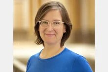 Person mit kurzen Haaren und Brille blickt lächelnd in die Kamera, Porträt vor unscharfem Hintergrund.