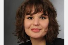 Person mit schulterlangen Haaren blickt direkt in die Kamera, Porträt vor neutralem Hintergrund.