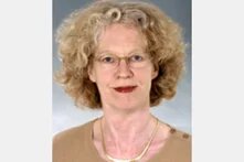 Foto: Porträt einer Person mit lockigem Haar und Brille vor grauem Hintergrund.