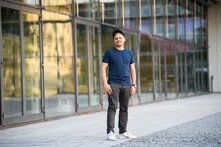 Eine Person mit kurzem Bart und Schnurrbart steht aufrecht vor einer Glasfassade, trägt Kappe, T-Shirt, Hose und Sneaker, blickt ernst zur Kamera.