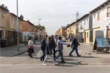 Das Foto zeigt eine Gruppe von Menschen, die eine Straße in einem südafrikanischen Township überquert, was ein typisches städtisches Wohngebiet am Rande von Städten oder Gemeinden darstellt. 