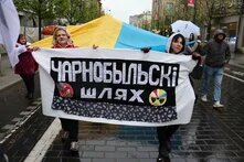 Zwei Frauen halten bei Regen auf einer Straße ein Banner mit kyrillischer Aufschrift, während hinter ihnen Menschen unter einem großen blau-gelben Tuch und mit Regenschirmen gehen.