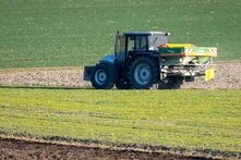Tractor with rear spreader drives across field, dispersing material; layered farmland stretches into background.