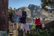 Laundry hangs on lines in a courtyard with brick walls and plants; a large mountain rises in the background under a clear sky.