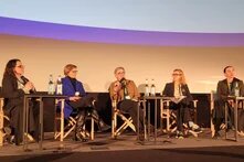 Five people sit on stage at a panel discussion. The person in the center speaks into a microphone; small tables with water bottles in front.