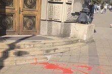 Red paint on ground in front of building steps; door and ornate facade left, pedestrians in background right.