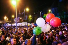 Dichte Menschenmenge bei Nacht auf Straße, viele stehen eng. Person hält Ballons. Lichter, Zelt und Gebäude im Hintergrund.