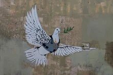 Graffiti einer fliegenden Taube mit Olivenzweig, auf Wand gemalt; Zielkreuz auf ihrem Körper, daneben kleiner Kasten an der Wand.