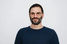 Mann mit Brille und Bart blickt lächelnd in die Kamera, neutraler Hintergrund.