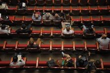 People sit scattered across rows of red seats in an auditorium, some reading or using phones and tablets.