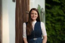 Frau in Tracht lehnt an Holzwand und lächelt in die Kamera, im Hintergrund Grünpflanzen.