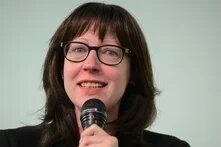 Frau mit Brille spricht lächelnd in ein Mikrofon vor hellem Hintergrund.