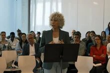 Frau mit Locken spricht an Rednerpult vor sitzendem Publikum in hellem Raum.