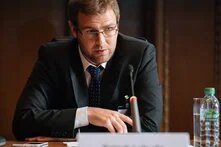 Mann mit Brille spricht an Konferenztisch ins Mikrofon, vor ihm Namensschild und Wasserflasche.