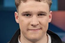 Junger Mann mit lockigen Haaren vor blauem Hintergrund, leicht lächelnd.