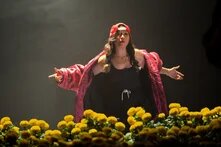 Person in red cap and open jacket stands on stage behind yellow flowers, arms spread wide.