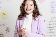 Lächelnde Person vor Whiteboard mit Notizen und Haftzetteln, gestikulierend im Gespräch.